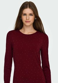 Vinröd långärmad topp med rund halsringning, prydd med utspridda blå och silverfärgade prickar för ett strukturerat utseende. Mjuk, stretchig tyg.