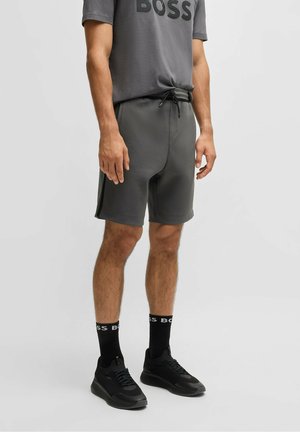 Mand iført grå shorts med snøre, grå T-shirt med teksten "BOSS", sorte sokker med "BOSS" logo og sorte sneakers, stående foran en ensfarvet baggrund.