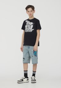 T-shirt grafico nero con testo bianco, abbinato a pantaloni bermuda di jeans blu chiaro con graffiti colorati e dettagli strappati. Sneakers bianco e nero.