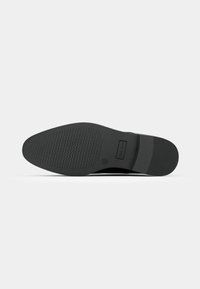Semelle de chaussure habillée noire avec motif de prise texturé et zone rectangulaire pour logo près de la voûte plantaire sur fond gris clair.