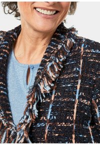 Strukturierte, mehrfarbige Strickjacke in Schwarz, Blau und Braun mit Fransenakzenten; über einem hellblauen Oberteil mit geknöpftem Ausschnitt getragen.