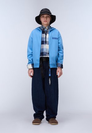 Nuori henkilö, jolla on musta lierihattu, sininen vetoketjullinen huppari ruutupaidan päällä, leveät tummat farkut ja ruskeat slip-on-kengät, seisomassa harmaan taustan edessä.