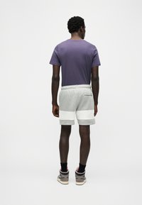 Άνδρας που φοράει μωβ μπλουζάκι, γκρι και λευκό σορτς, μαύρες κάλτσες και μπεζ και ροζ αθλητικά παπούτσια, όρθιος με την πλάτη προς τα εμάς σε λευκό φόντο.