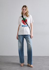 Maglietta bianca con grafica di fragole verdi e rosse e testo "BALLY". Abbinata a jeans blu sbiaditi a gamba larga e sandali beige con tacco a blocco.