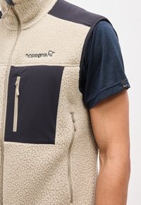 Μπεζ και μαύρο γιλέκο από φλις με εμπρόσθιο φερμουάρ και τσέπη στο στήθος φορεμένο πάνω από μπλε κοντομάνικο πουκάμισο σε άνω μέρος σώματος ατόμου.