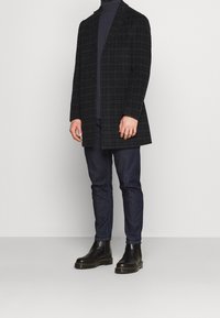 Rutig överrock i mörkgrön och svart, med enkelknäppning, kombinerad med en mörk polotröja, blå jeans och svarta ankelboots.