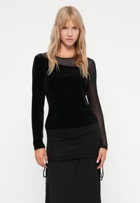 Black long-sleeve top with a velvet texture and sheer mesh neckline. Worn with a fitted black skirt featuring gathered detailing at the waist.