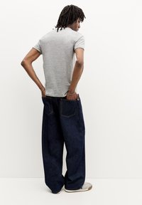 Tricou gri cu mânecă scurtă, asortat cu blugi rombiști de culoare albastru închis. Are buzunare mari pe spate și o croială lejeră. Fundal alb.