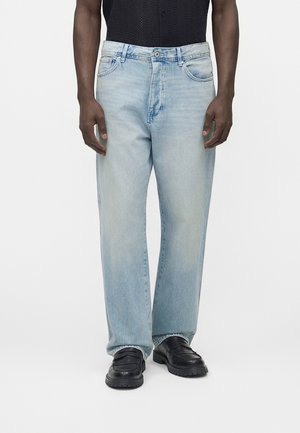 Mężczyzna ubrany w jasnoniebieskie dżinsy o prostych nogawkach, czarne mokasyny oraz czarną koszulę z krótkim rękawem, stojący na jednolitym tle.