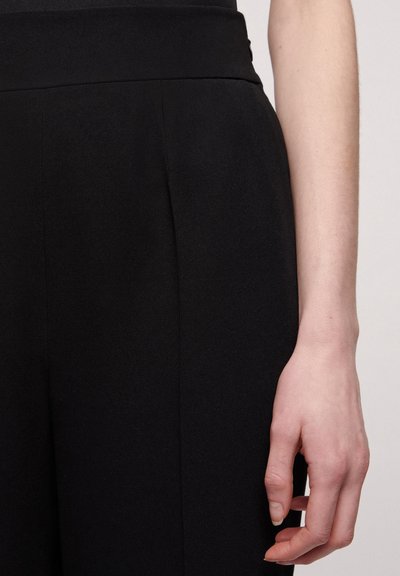 Pantalon noir avec une taille large, en tissu doux et des plis sur le devant, mains partiellement visibles près de la taille, sans détails visibles.