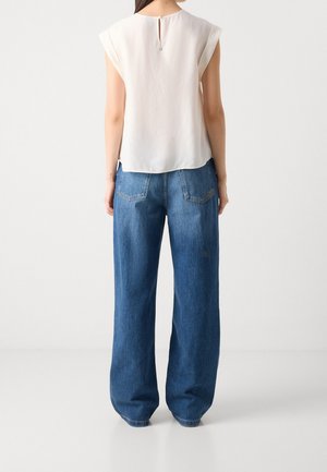 Kvinde iført en ærmeløs hvid bluse og løstsiddende blå jeans, der står med ryggen til kameraet mod en ensfarvet baggrund.