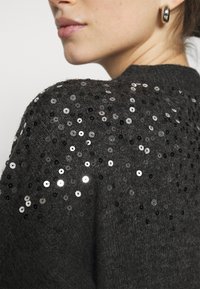 Gros plan sur une personne portant un pull en tricot gris foncé orné de paillettes noires et argentées brillantes sur l'épaule et le haut du dos.