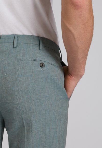 Close-up of a person’s waist wearing light blue dress pants with back button pocket and white shirt, hand in front pocket.