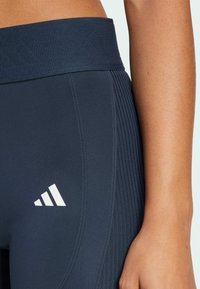 Marinen blå leggings med en tekstureret, ribbet talje. Har et hvidt Adidas-logo på den nederste venstre side. Glat stof med lodrette striber.
