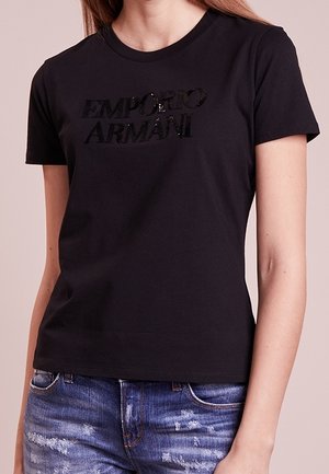Kvinde iført en sort Emporio Armani t-shirt med korte ærmer og blå slidte jeans, vist fra nakke til hofter.