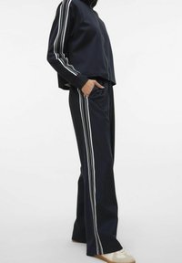 Marineblauw trainingspak met witte zijkanten, bestaande uit een jack met rits en een relaxte broek. Zachte stof met een gladde textuur.