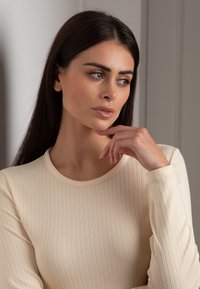 Top a maniche lunghe, in maglia ribbed di colore crema, con scollatura rotonda e silhouette aderente, accompagnato da capelli lisci e dritti e trucco naturale.