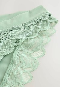 Muntgroene stof met ingewikkelde kanten rand met golvende randen en bloemmotieven. Zachte textuur met delicate afwerking.