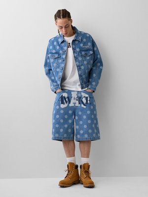 Personne avec des cheveux tressés portant une veste en jean bleue à pois et un short avec le texte « AWAKE », des chaussettes blanches et des bottes marron à lacets.