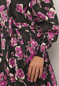 Blumenkleid aus schwarzem Stoff mit pinken, weißen und lila Blumen. Enthält einen Taillengürtel und geraffte Manschetten an den langen Ärmeln.