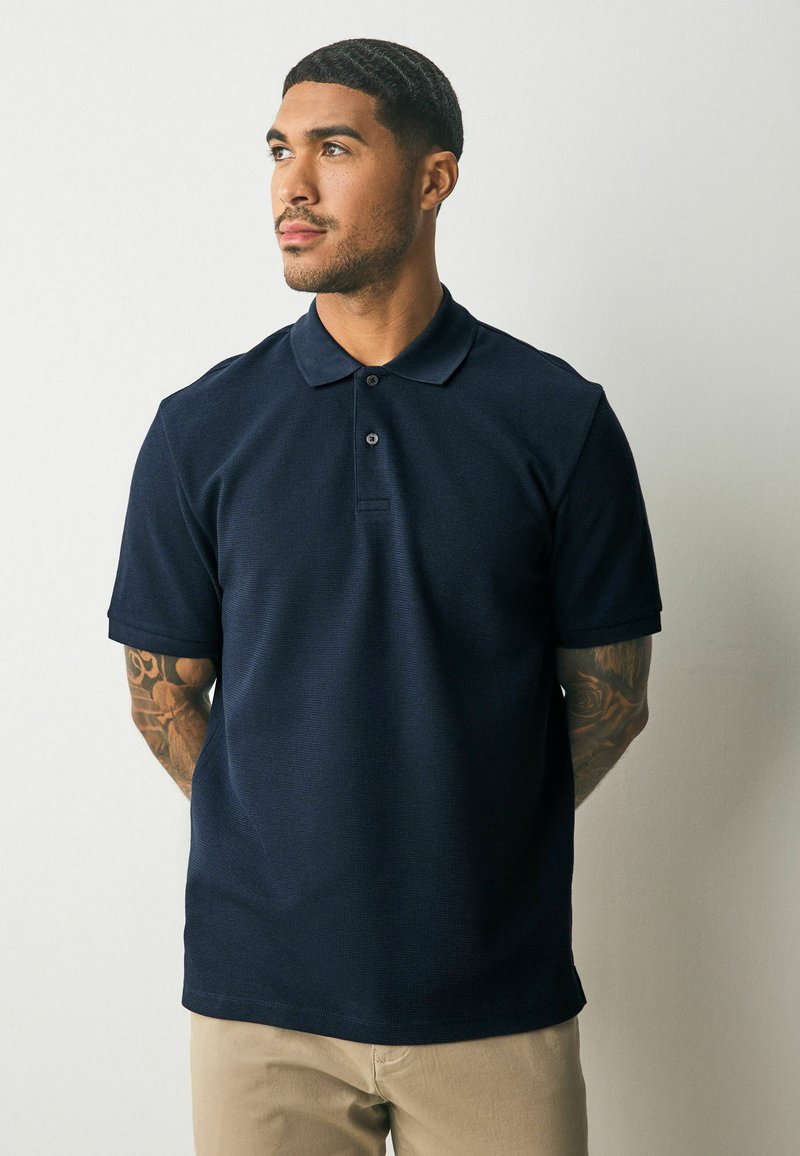 Marineblå polo t-shirt med korte ærmer, struktureret stof, to-knaps lukning og spids krave. Båret med khaki bukser.