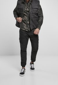 Černá bezrukávová vesta s několika kapsami, nošená přes maskáčovou košili s knoflíky. Černé kalhoty typu cargo a černé tenisky doplňují vzhled.