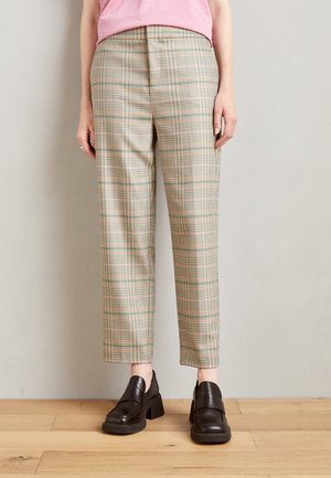Pantalon à carreaux en beige clair, vert et rose doux, avec une coupe ajustée et une jambe raccourcie. Porté avec des mocassins noirs sur un sol en bois.