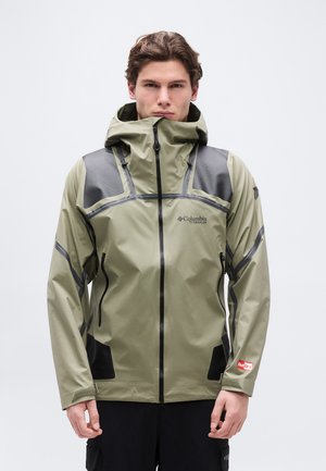 Giovane uomo che indossa una giacca impermeabile verde Columbia Titanium con cappuccio alzato, in piedi contro uno sfondo chiaro e uniforme.