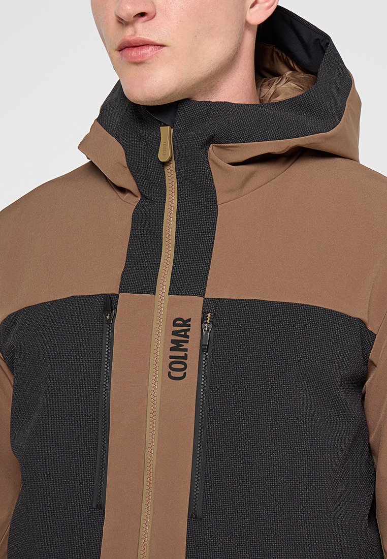 Veste à capuche marron et noire avec un tissu texturé et des sections lisses, dotée d'une fermeture éclair à l'avant et de deux poches zippées. Logo "COLMAR" visible.