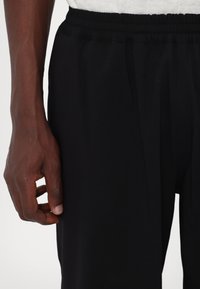 Sorteidrettsbukser med elastisk liv, laget av et glatt, lett stoff og med innsnevring. Hånd som hviler på sideleden.