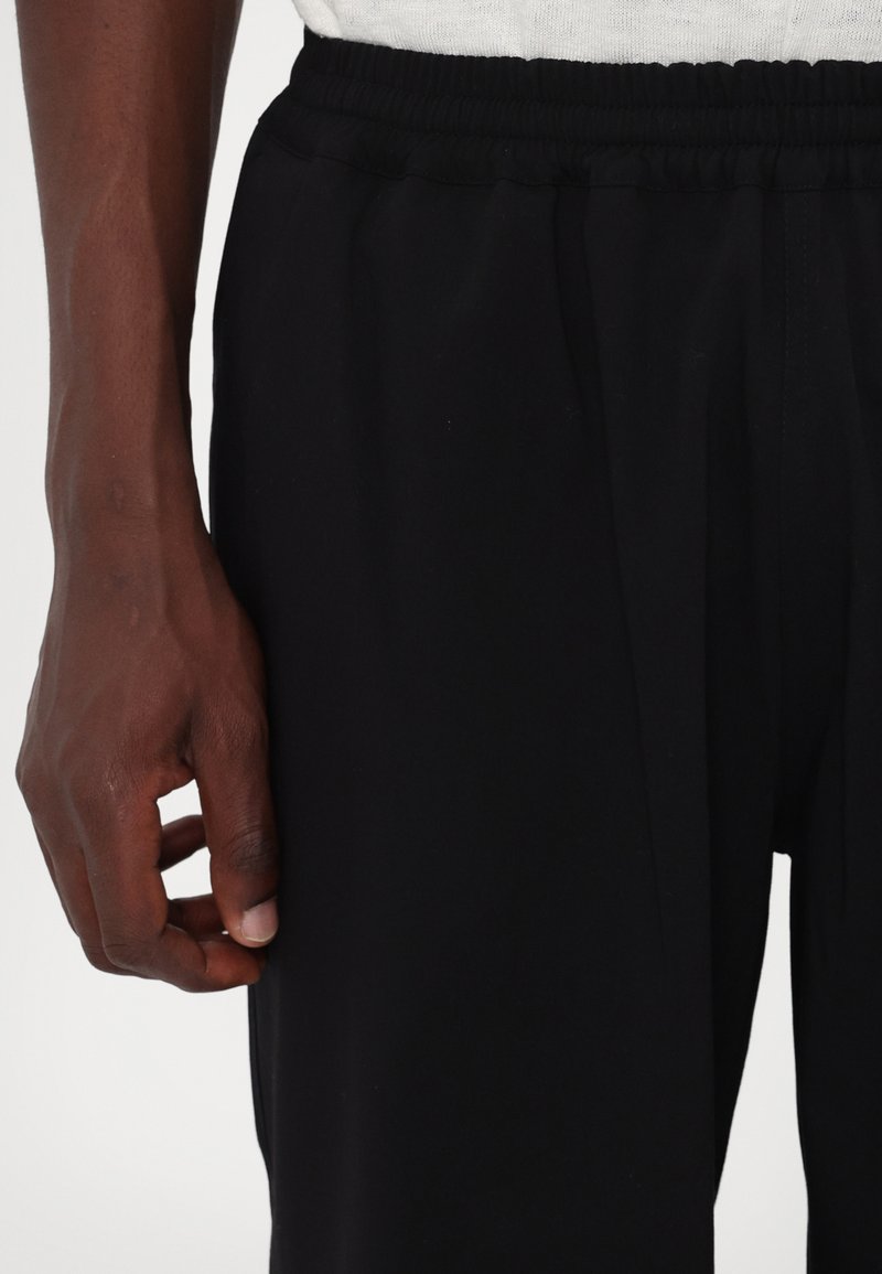 Pantaloni sportivi neri con vita elastica, realizzati in un tessuto leggero e liscio con un design affusolato. Mano appoggiata sulla cucitura laterale.