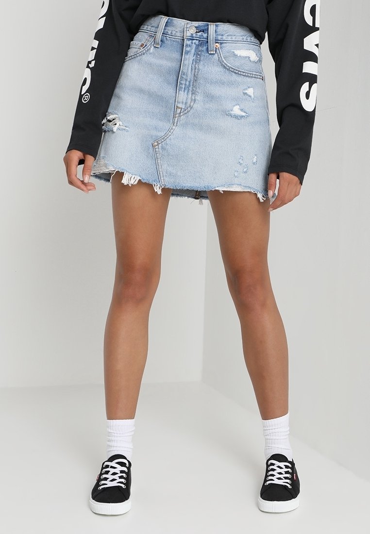 Femme portant une mini-jupe en denim déchirée bleu clair, un haut noir à manches longues, des chaussettes blanches et des baskets noires, se tenant contre un fond blanc.