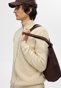 Beiger Zip-Hoodie aus strukturiertem Stoff, kombiniert mit einer braunen Umhängetasche. Eine braune Mütze rundet den Look ab.