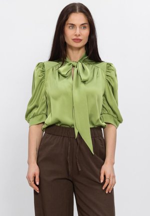 Femme aux longs cheveux bruns portant un chemisier en satin vert clair à manches bouffantes et un grand nœud papillon au col, associé à un pantalon marron.