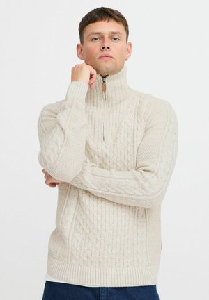 Suéter de punto trenzado color crema con un cuello alto, que cuenta con una cremallera y puños y dobladillo acanalados. Textura suave con patrones intrincados.