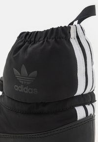 Černá voděodolná bota s nařaseným horním lemem, třemi bílými pruhy a tónovaným logem. Vyrobená z hladké látky a gumové podrážky.