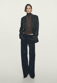 Blazer nero sopra un dolcevita marrone e pantaloni larghi scuri. Collana visibile. Tessuti lisci con elementi di design su misura.