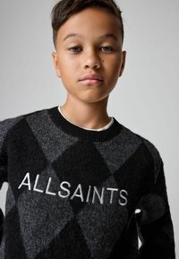 Ragazzo con capelli corti che indossa un maglione a diamante bianco e grigio con "ALLSAINTS" ricamato sul petto, sfondo neutro.