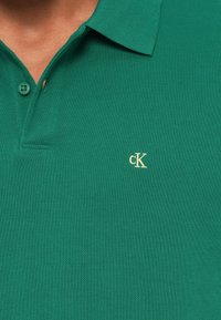 Πράσινο πόλο πουκάμισο κατασκευασμένο από υφασμάτινη υφή, με πατιλέτα και δύο κουμπιά καθώς και ένα μικρό, κεντημένο χρυσό λογότυπο στο στήθος.