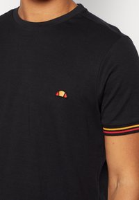 Black cotton t-shirt with a ribbed crew neckline and short sleeves. Features a small embroidered logo in yellow, red, and orange on the chest.