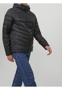 Hombre con chaqueta acolchada negra con capucha y jeans azules caminando contra un fondo claro liso.