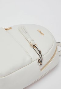 Sac à dos en similicuir blanc de forme arrondie, avec des accents dorés, un détail en pampille et une fermeture éclair. Logo embossé à l'avant.