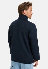 Navy fleece jacket with a high collar, textured fabric, and a relaxed fit. Features a seam across the shoulders and long sleeves.