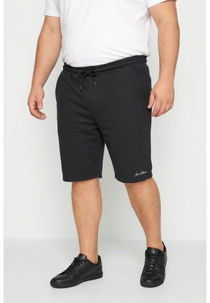 Uomo con pantaloncini neri con coulisse, maglietta bianca e scarpe da ginnastica nere, in piedi davanti a uno sfondo chiaro e uniforme.