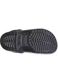 Black rubber Crocs bottom sole with textured tread pattern, featuring size markings and a small logo. Accented with a blue edge design.