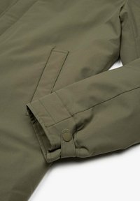 Primo piano di una manica di giacca verde oliva con polsino regolabile a bottone a pressione e una tasca laterale con apertura verticale.