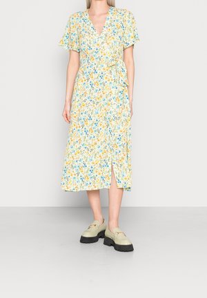 Femme portant une robe portefeuille fleurie mi-longue à manches courtes et des mocassins à plateforme beiges, debout devant un fond clair uni.