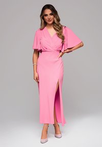 Różowa sukienka do ziemi z krótkimi, rozszerzanymi rękawami, głębokim dekoltem w kształcie litery V i rozcięciem sięgającym do uda, uszyta z gładkiego materiału. W zestawie z jasnoróżowymi szpilkami.