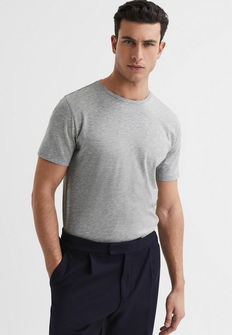 Tricou gri cu mânecă scurtă, realizat dintr-un material moale, având un guler rotund și un design slim-fit, asortat cu pantaloni întunecați. Fără modele sau accente vizibile.