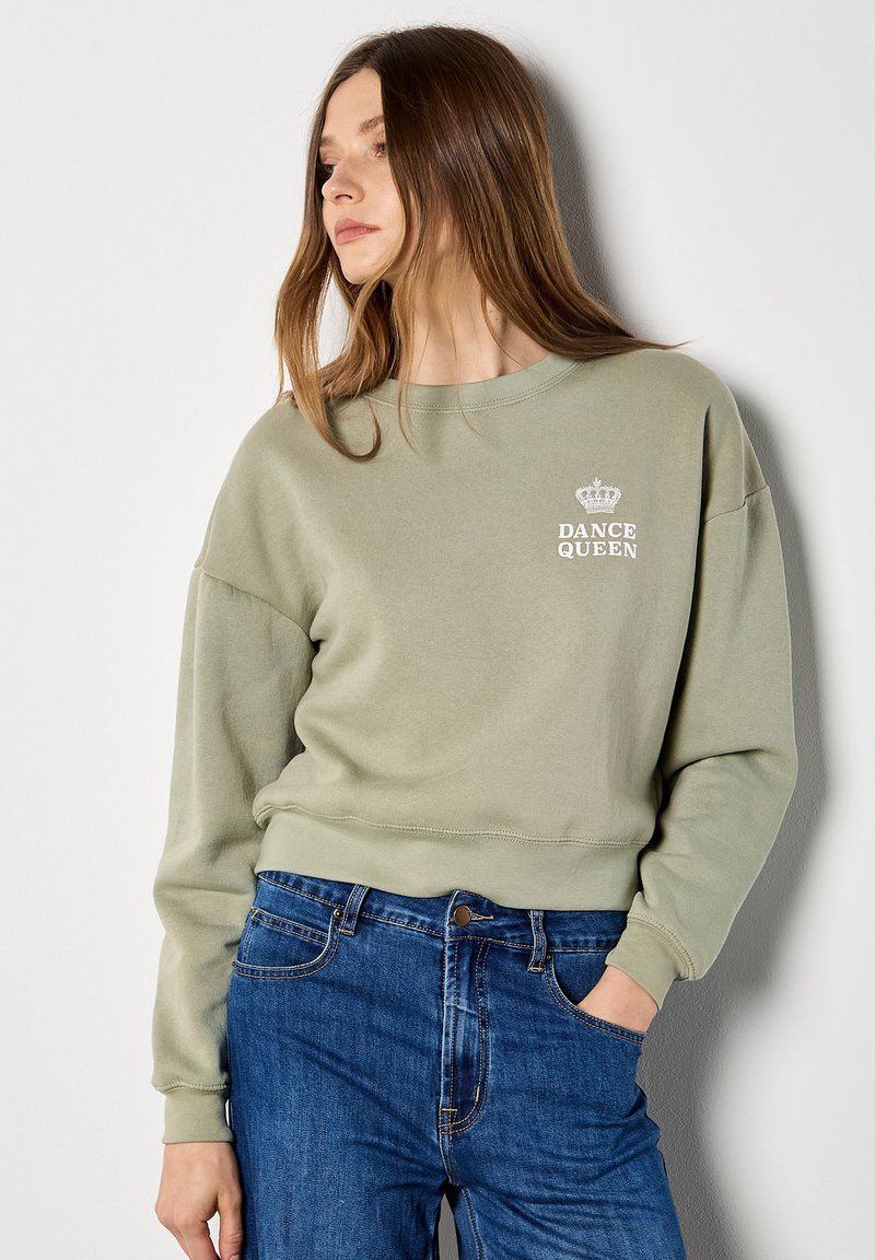 Femme en sweat-shirt vert clair avec le texte « Dance Queen » et un motif de couronne, portant un jean bleu, debout contre un mur blanc, regardant à gauche.